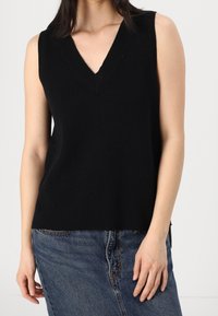 Femme portant un haut tricoté noir sans manches à col en V, associé à une jupe en jean bleue, vue des épaules aux hanches sur un fond uni.