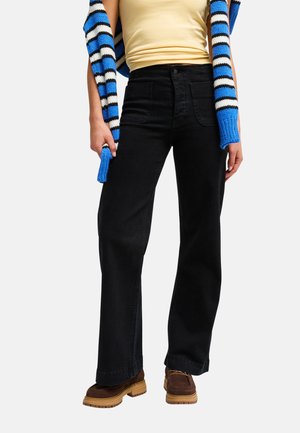 Personne portant un pantalon noir taille haute à jambes larges, des chaussures marron, un haut jaune et un pull rayé bleu, blanc et noir sur les épaules.