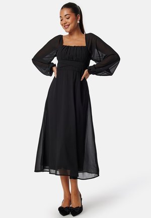 Abito maxi nero con lunghe maniche a sbuffo trasparenti, scollatura quadrata e dettagli arricciati sul corpetto. Tessuto fluente con una leggera lucentezza.