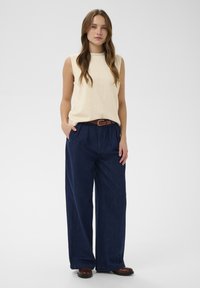 Bežinis be rankovių topas, derinamas su tamsiai mėlynomis plačiomis kelnėmis su raukiniu priekyje ir rudos spalvos diržu, sukuriantys atsipalaidavusį siluetą.