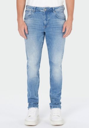 Jean slim bleu clair porté avec des baskets blanches et une chemise blanche, présenté de la taille aux pieds sur un fond uni.
