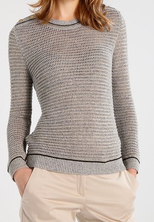 Kobieta w szarym swetrze o fakturze dzianiny z złotymi guzikami na ramionach i beżowych spodniach, z rękami w kieszeniach, na jednolitym tle.