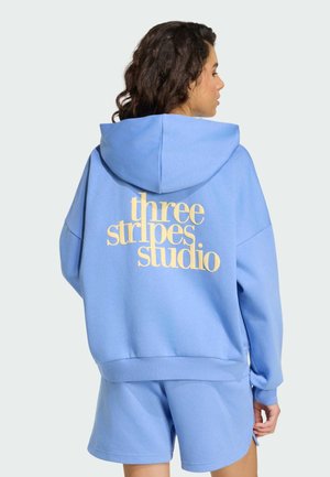 Femme aux cheveux bouclés foncés portant un sweat à capuche bleu clair et un short assorti, avec au dos du sweat à capuche un texte jaune "three stripes studio."