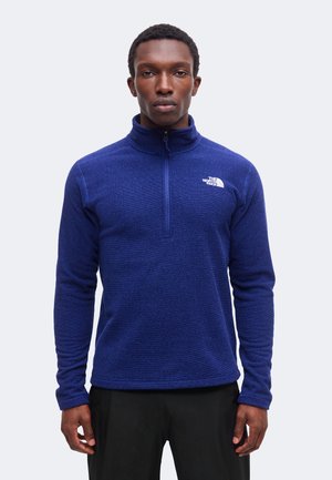Homme portant un polaire bleu à manches longues avec une fermeture éclair au quart et le logo The North Face sur la poitrine, debout devant un fond uni.