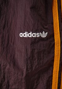 Adidas treniruočių striukė tamsiai violetinės spalvos audiniu su tekstūruotu paviršiumi, su baltu logotipu ir oranžiniais tinkliniais akcentais šonuose.