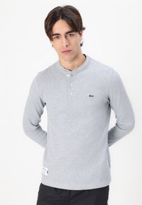 Siv gray majica s dolgimi rokavi henley z teksturiranim vzorcem, ki ima zapenjanje na tri gumbe in majhen zeleni logotip na levem prsnem delu.