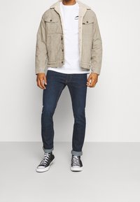Ljus beige jeansjacka med fuskpälskrage, vit t-shirt, mörkblå jeans och svarta höga sneakers med vita sidor.