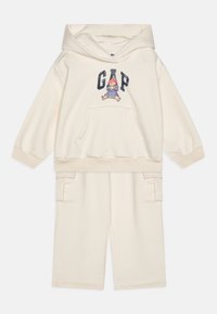 Set de hanorac și pantaloni scurți de culoare crem deschis, realizat dintr-un material moale. Dispune de un buzunar frontal, manșete ribbate și o grafică cu un personaj pe hanorac.