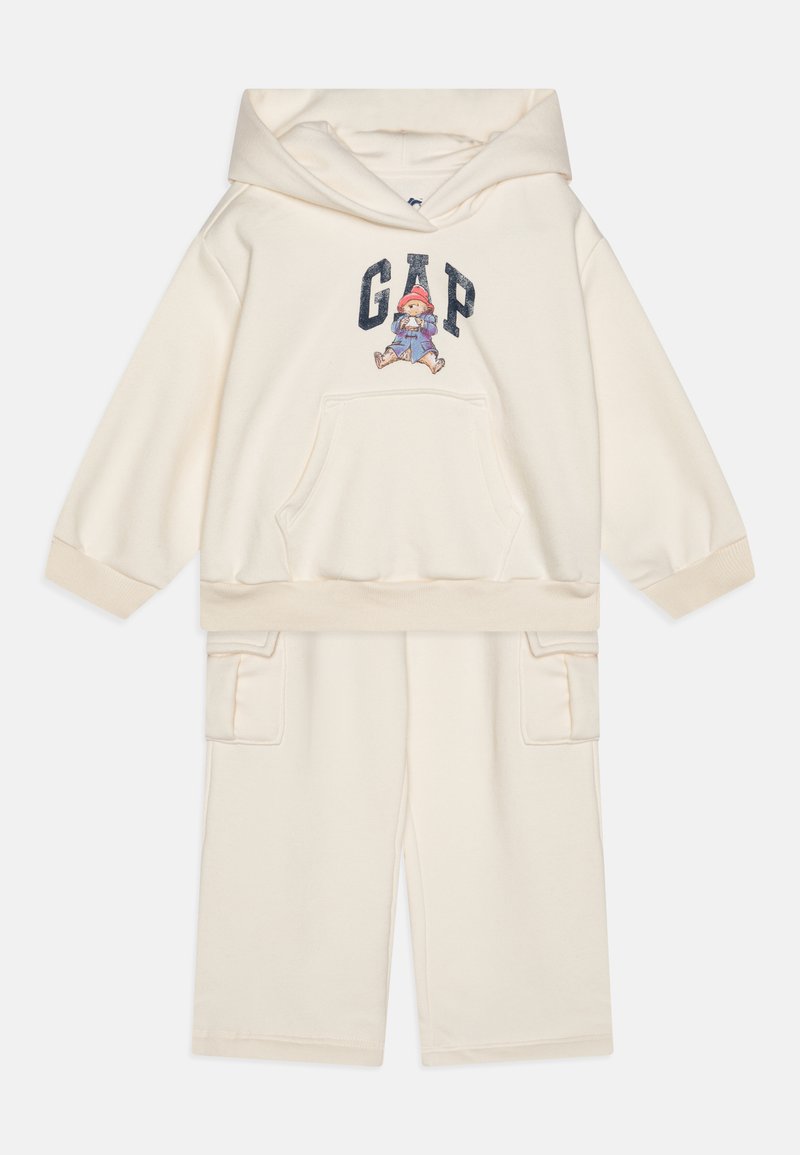 Set de hanorac și pantaloni scurți de culoare crem deschis, realizat dintr-un material moale. Dispune de un buzunar frontal, manșete ribbate și o grafică cu un personaj pe hanorac.