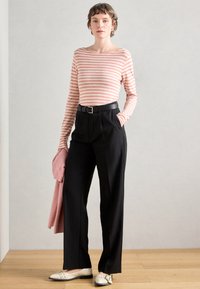 Camisa de mangas longas às riscas em rosa e branco, combinada com calças pretas de cintura alta e sapatos brancos. O modelo segura um suéter rosa.