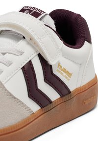 Hvide sneakers med maroon detaljer, der har en hook-and-loop rem, struktureret suede og en gum-sål. Guld branding læser "hummel handball perfekt."