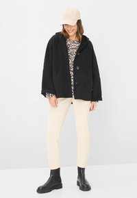 Sort oversized jakke med knapper, beige mønstret top, cremefarvede fløjlsbukser og sorte ankelstøvler. En beige cap fuldender looket.