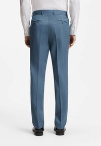 Homme portant un pantalon de costume bleu sur mesure, des chaussures de ville noires et une chemise blanche à manches longues, vu de dos sur un fond blanc.