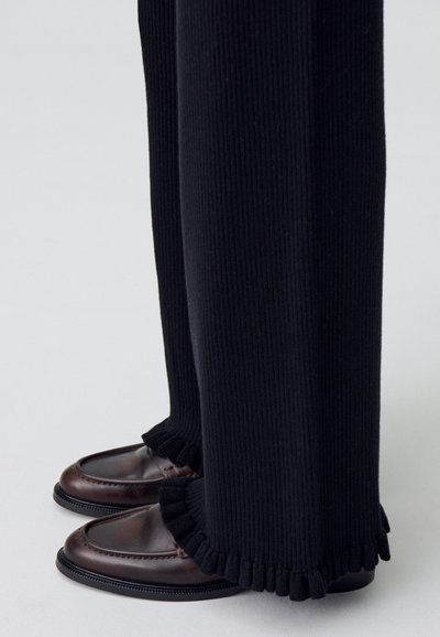 Pantalones negros de canalé con puños deshilachados, combinados con mocasines de cuero marrón que tienen una punta redondeada y un detalle de costura decorativa.