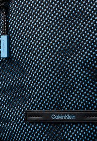 Tejido de malla negra con respaldo azul, que presenta una etiqueta de cuero negro grabada con "Calvin Klein". Incluye un tirador de cremallera azul.