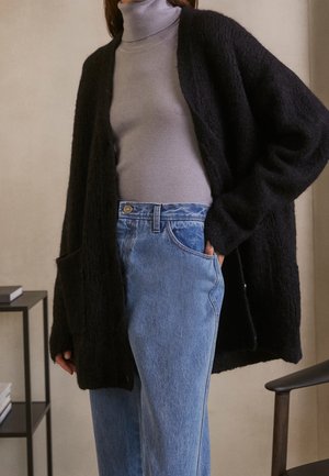 Sort fuzzy cardigan med åben front, båret over en lys grå turtleneck, parret med lyseblå lige ben jeans. Simpel lomme-detalje synlig.