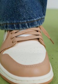 Baskets blanches et beiges avec des lacets beiges, portées avec un jean en denim bleu sur une surface verte.