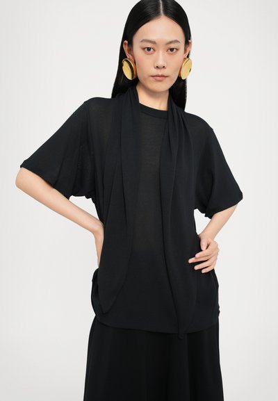 Top nero a maniche corte, con un design drappeggiato sul davanti e una texture morbida. Abbinato a grandi orecchini circolari dorati.
