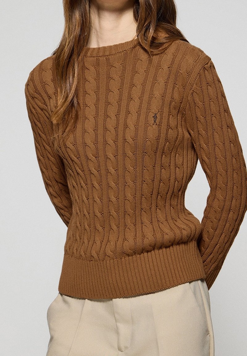 Pull marron en maille torsadée avec ourlet côtelé porté avec un pantalon beige, visible du milieu de la poitrine jusqu'aux hanches.