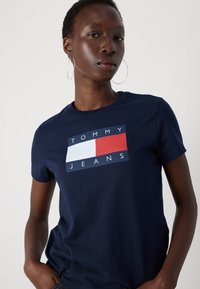 Námornícky modré bavlnené tričko s grafickým dizajnom a textom "TOMMY JEANS" a červenými, bielymi a modrými obdĺžnikovými akcentmi. Krátke rukávy.