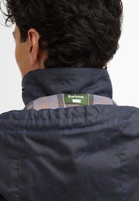Casaco azul marinho com gola alta, apresentando um detalhe xadrez e um rótulo verde da Barbour na gola. Textura suave e design estruturado.