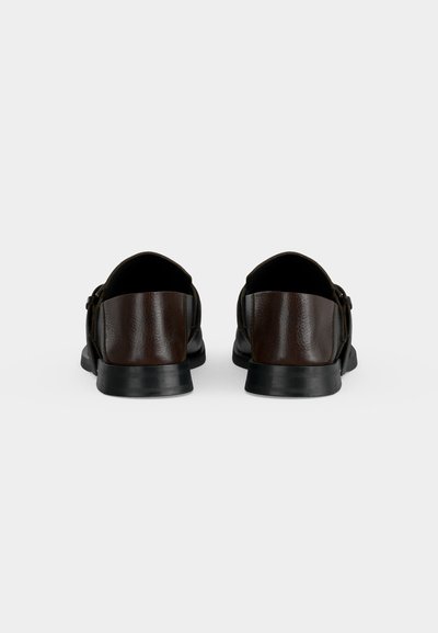 Paire de chaussures habillées en cuir brun foncé vues de dos, avec semelles noires et petits talons sur un fond clair uni.