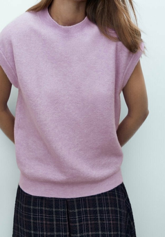 Pull sans manches rose clair en maille avec un col rond et un ourlet côtelé, associé à une jupe à carreaux foncés, présentant une texture lisse.