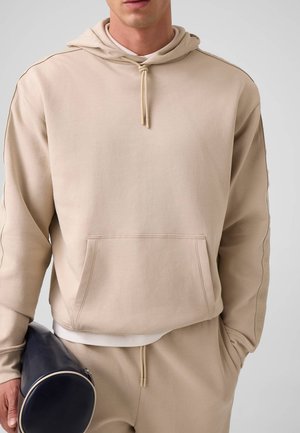 Mann in beigefarbenem Hoodie und Jogginghose hält eine navyfarbene zylindrische Tasche. Nur sein Oberkörper und ein Teil seines Gesichts sind vor neutralem Hintergrund sichtbar.