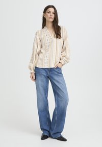 Blouse à rayures blanches et beiges avec manches bouffantes, ornée de motifs en losange. Associée à un jean large bleu clair et des chaussures noires à bout pointu.