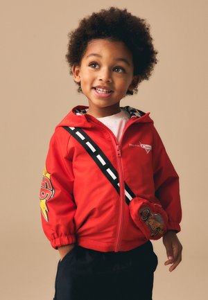 Junges Kind mit lockigem Haar lächelt, trägt eine rote Jacke mit Reißverschluss und Cars-Motiven sowie eine kleine Figurentasche quer über der Brust.