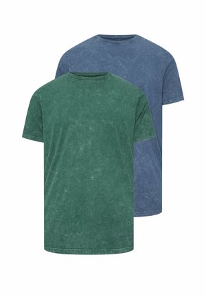 Mężczyzna w niebieskiej koszuli z krótkim rękawem poprawiający swój rękaw, stojący na tle jednolitego białego tła.