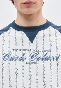 Κοντινό πλάνο ενός ατόμου που φοράει λευκό και μπλε πουκάμισο με κάθετη γραφή και μεγάλο κείμενο που λέει "Το να κερδίζεις δεν φαινόταν ποτέ καλύτερο Carlo Colucci Est. 1978".