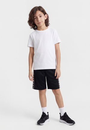 Enfant aux cheveux mi-longs portant un t-shirt blanc, un short de sport noir avec logo sur le côté, des chaussettes blanches et des baskets noires, debout devant un fond uni.