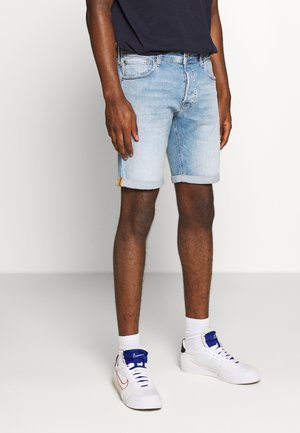 Homme portant un short en denim bleu clair, des baskets blanches avec des touches bleues, des chaussettes blanches et un t-shirt bleu marine foncé, debout devant un fond blanc.