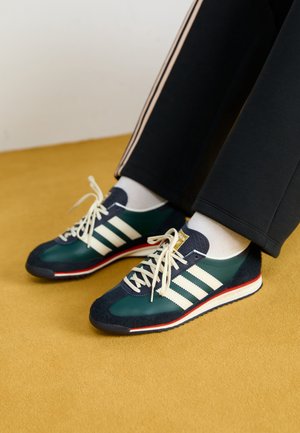 Persoon die donkerblauwe en groene sneakers met witte strepen draagt, witte sokken en zwarte broek met beige zijstrepen op een gele vloer.