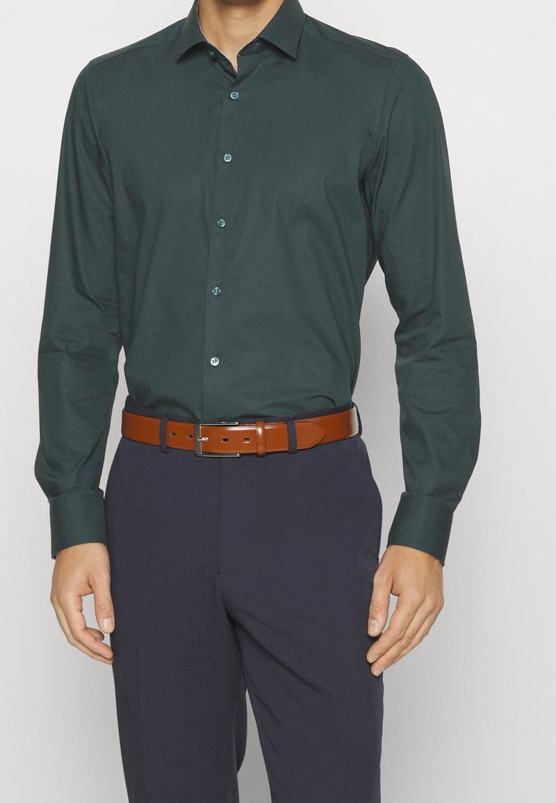 Homme portant une chemise à manches longues boutonnée vert foncé rentrée dans un pantalon sombre avec une ceinture en cuir marron.