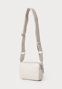 Beige rechteckige Handtasche mit glatter, glänzender Oberfläche und einem strukturierten Tragegurt, auf dem "Calvin Klein" in einem wiederholenden Muster zu sehen ist, silberne Beschläge.