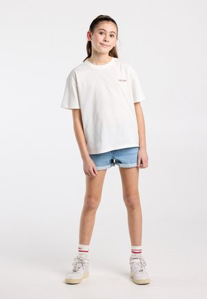 Young girl standing in white T-shirt, rolled denim shorts, white sneakers, and striped socks against plain background.