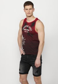 Prúžkovaná tank top v červenej a čiernej farbe s textom a grafikou palmových stromov; nosená s čiernymi denimsovými šortkami s opotrebovaným vzhľadom.