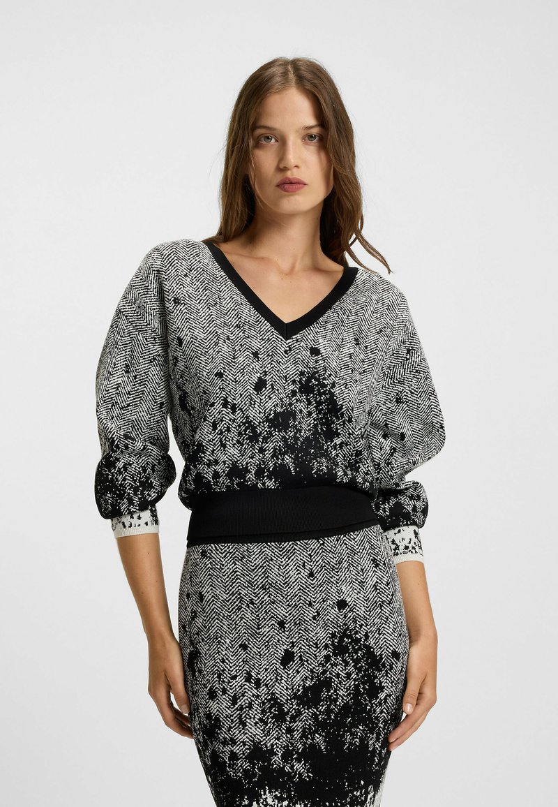 Schwarzer undweißer Strickpullover mit V-Ausschnitt, Hahnentrittmuster, puffierten Ärmeln und einem gerippten Bündchen. Strukturierter Stoff mit Spritzakzenten.