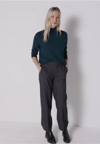 Dunkelgrüner, langärmeliger Pullover mit hohem Kragen, kombiniert mit hellgrauen, hochgeschnittenen Hosen. Das Model trägt schwarze Stiefeletten.