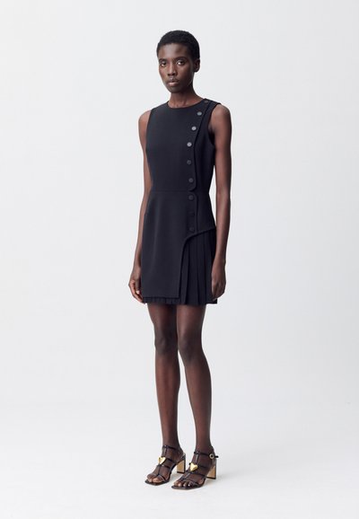 Una modella indossa un vestito nero senza maniche con bottoni e pieghe, abbinato a tacchi neri con cinturini, di fronte a uno sfondo neutro.