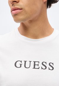 Νεαρός άνδρας που φοράει λευκό T-shirt με μαύρο λογότυπο "GUESS", ορατό από τον λαιμό έως το πάνω μέρος του στήθους, πάνω σε απλό φωτεινό φόντο.