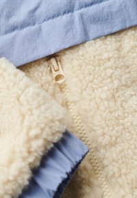Giacca in pile color crema con un interno morbido e texture. Presenta un colletto in tessuto blu chiaro e una cerniera beige con dettagli di branding.