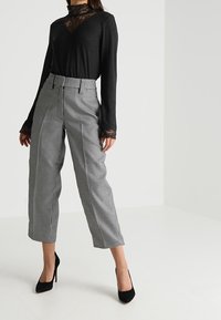 Woman wearing high-waist black-and-white checkered cropped pants, black lace long-sleeve top, and black high heels, standing in neutral background.