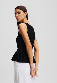 Juodas peplum stilius viršus su prigludusia liemene, be rankovių dizainu ir tekstūruotu audiniu. Modelis dėvi jį su baltais pantalonais, pabrėždamas švarią siluetą.