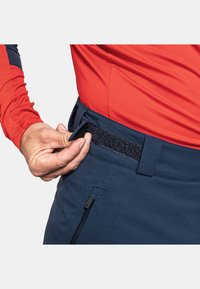 Blaue Hose mit Klettverschluss zur Taillenverstellung, Hand, die den Riemen greift, zeigt eine glatte Stoffstruktur und seitliche Reißverschlussdetails.