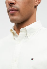 Camisa branca com botões, apresentando um pequeno logotipo bordado em vermelho, branco e azul. O tecido parece suave com um design de gola clássica.