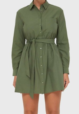 Femme portant une robe-chemise vert olive à manches longues avec des boutons sur le devant, une poche poitrine et une ceinture nouée à la taille.