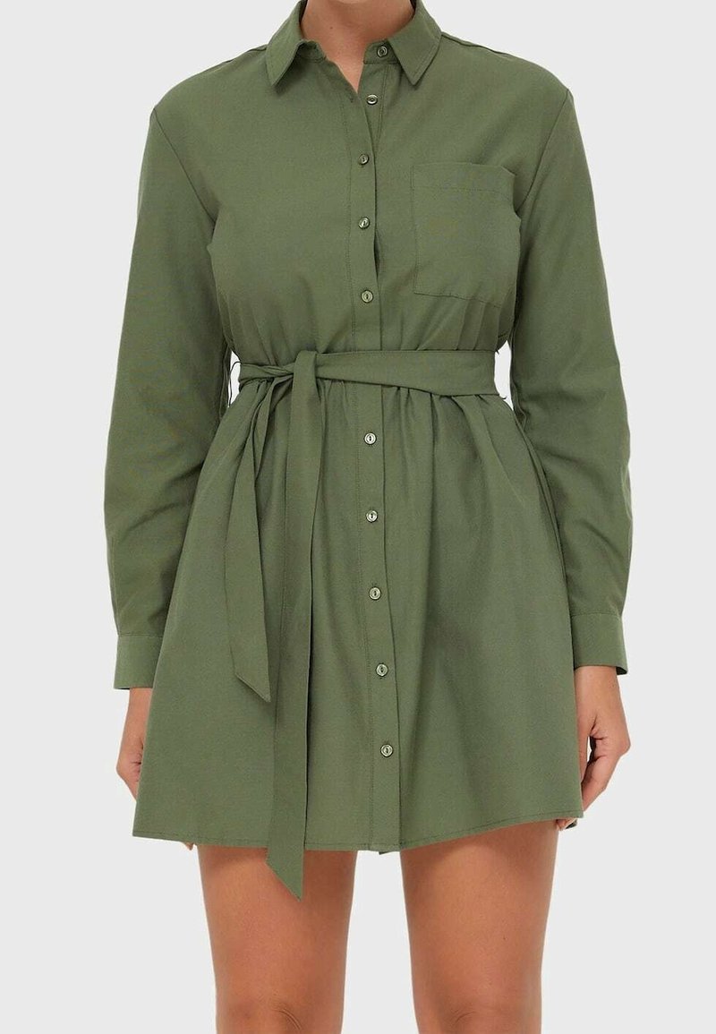 Femme portant une robe-chemise vert olive à manches longues avec des boutons sur le devant, une poche poitrine et une ceinture nouée à la taille.